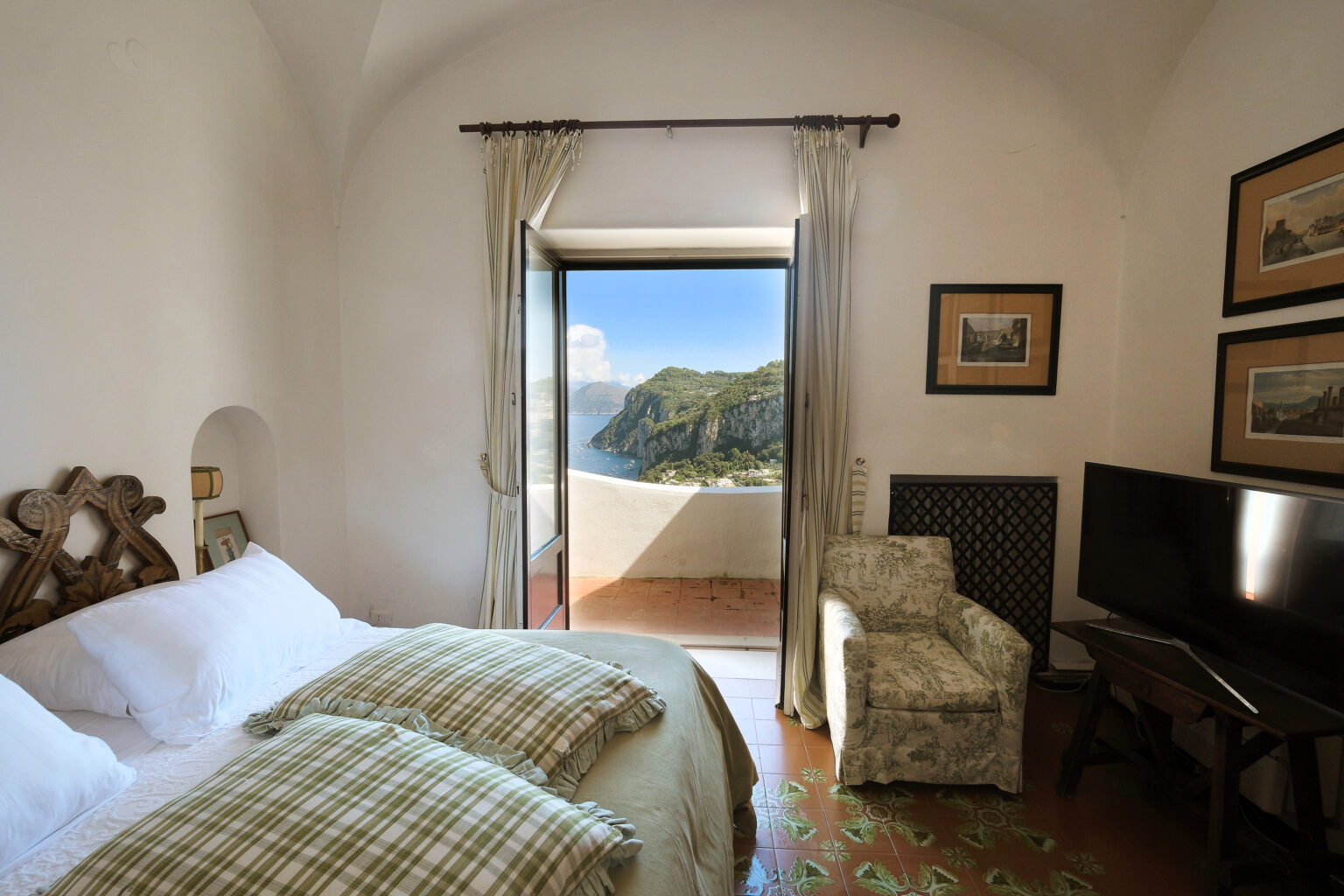 Cozy bedroom with two beds, decorative pillows, and a scenic view of mountains through an open door. Features a small armchair and a television, complemented by soft natural lighting and elegant decor. | Only on SopranoVillas