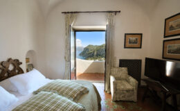 Cozy bedroom with two beds, decorative pillows, and a scenic view of mountains through an open door. Features a small armchair and a television, complemented by soft natural lighting and elegant decor.