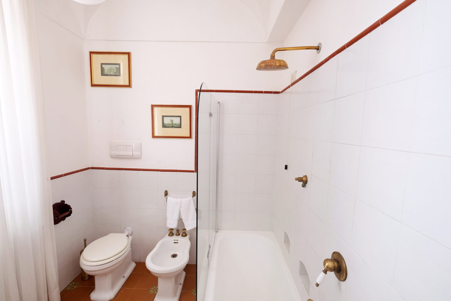 Modern bathroom featuring a bathtub, toilet, bidet, and shower, with white tiled walls and decorative artwork. Natural light filters through a sheer curtain, enhancing the clean and spacious atmosphere. | Only on SopranoVillas
