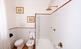 Modern bathroom featuring a bathtub, toilet, bidet, and shower, with white tiled walls and decorative artwork. Natural light filters through a sheer curtain, enhancing the clean and spacious atmosphere.