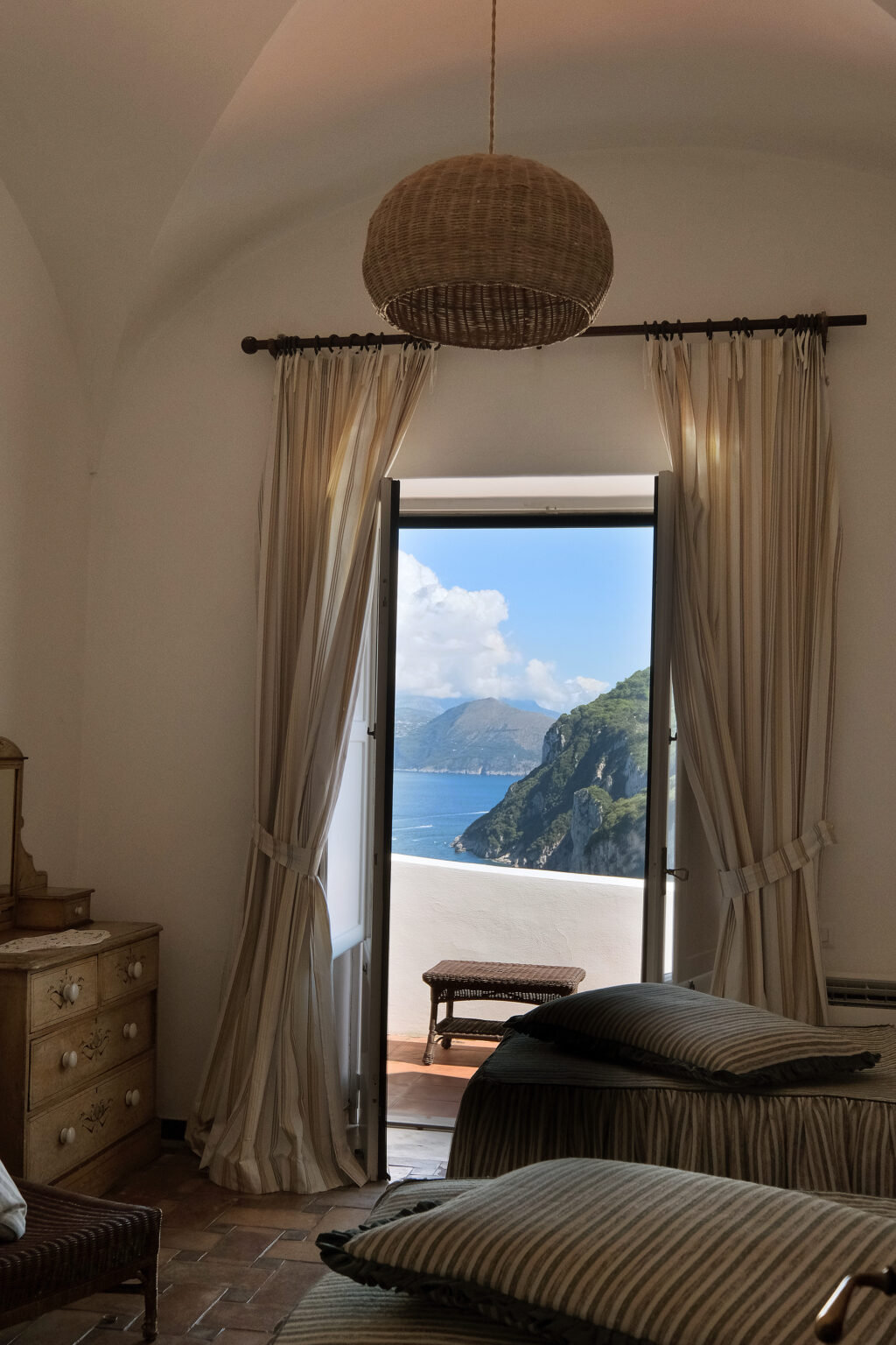 View from a stylish interior window showcasing a scenic coastal landscape with mountains and blue waters, framed by elegant curtains. | Only on SopranoVillas