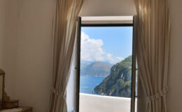 View from a stylish interior window showcasing a scenic coastal landscape with mountains and blue waters, framed by elegant curtains.