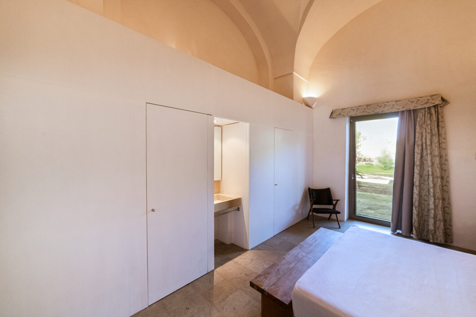 Modern bedroom interior featuring a minimalist design, with white walls, a wooden bed frame, and a chair. The room includes a doorway leading to a bathroom and large windows allowing natural light and garden views. | Only on SopranoVillas