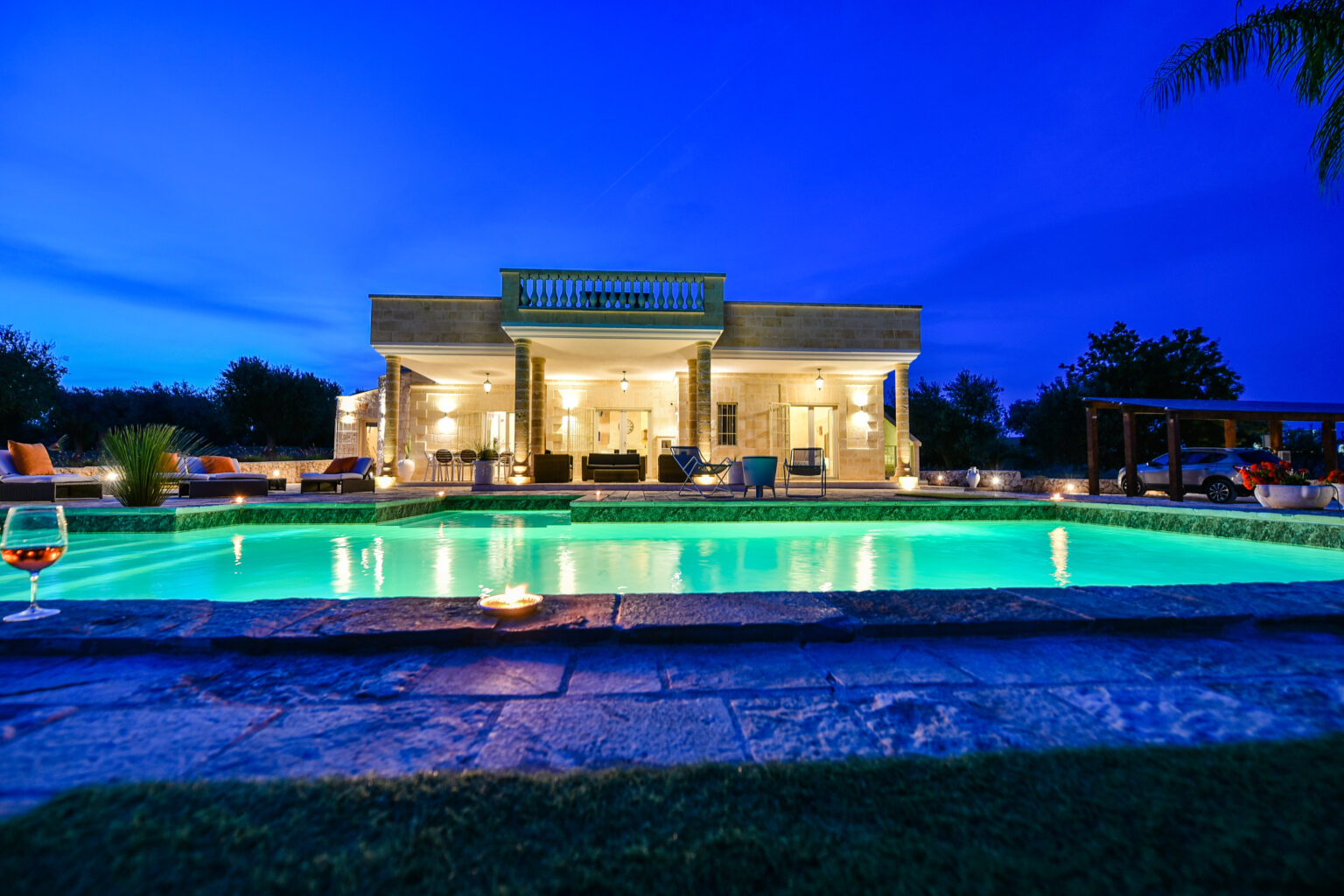 Luxury villa with illuminated pool at dusk, surrounded by lush greenery, showcasing outdoor seating and elegant architecture. Ideal for vacation rentals or luxury retreats. | Only on SopranoVillas