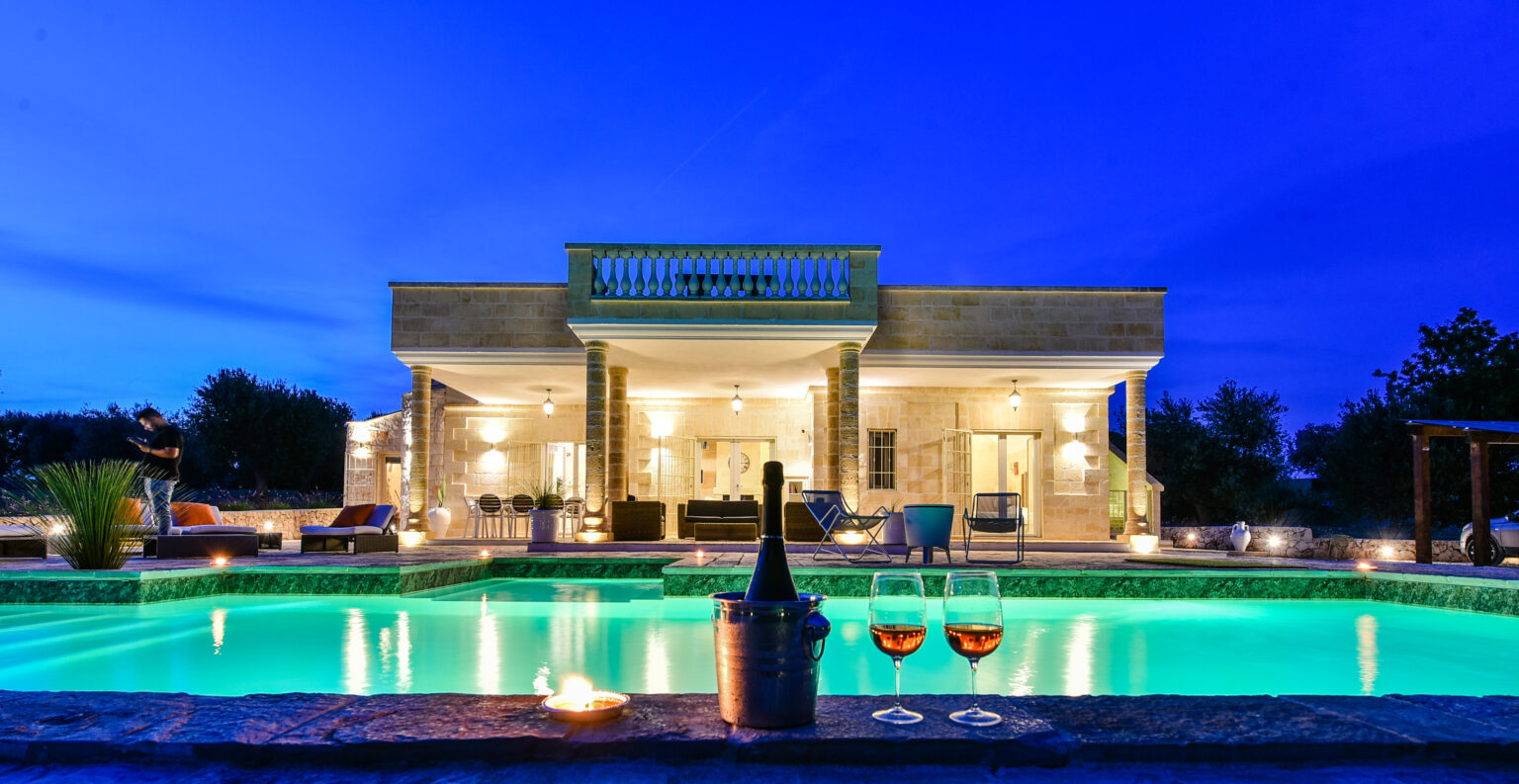 Luxury villa at dusk with a sparkling pool, featuring a bottle of champagne and two glasses in the foreground, highlighting an inviting outdoor living space and elegant architecture. | Only on SopranoVillas
