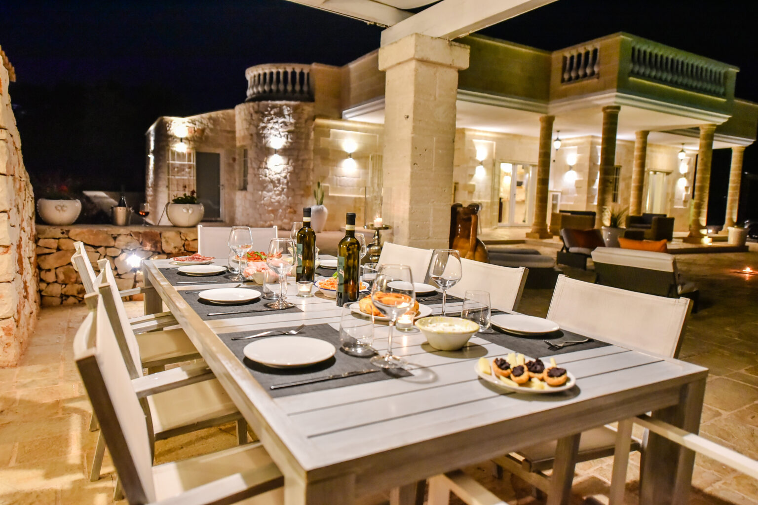 Elegant outdoor dining setup at night featuring a beautifully arranged table with plates, glasses, and gourmet desserts, surrounded by a luxurious villa with modern architecture and ambient lighting. | Only on SopranoVillas