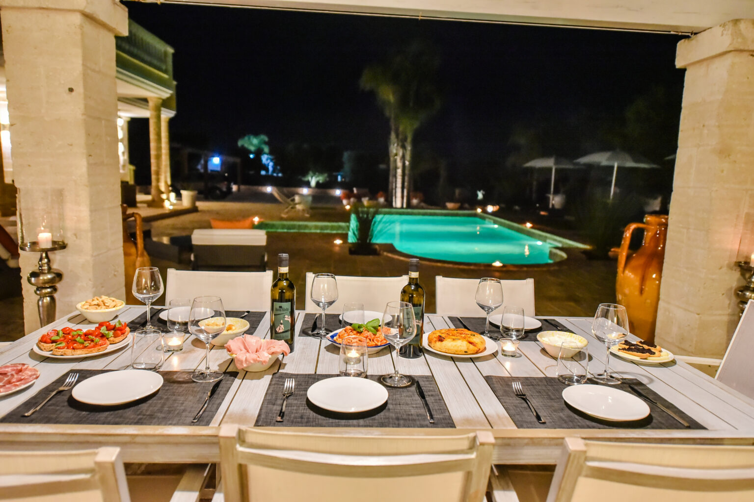 Elegant outdoor dining setup at night featuring a beautifully arranged table with a variety of gourmet dishes, wine bottles, and glasses, overlooking a serene illuminated pool and lush greenery. | Only on SopranoVillas