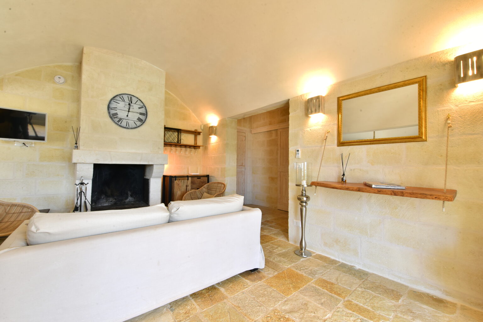 Cozy living room featuring a white sofa, stone fireplace, and natural light from wall sconces. The decor includes a wall clock and a wooden shelf, creating a warm and inviting atmosphere. | Only on SopranoVillas