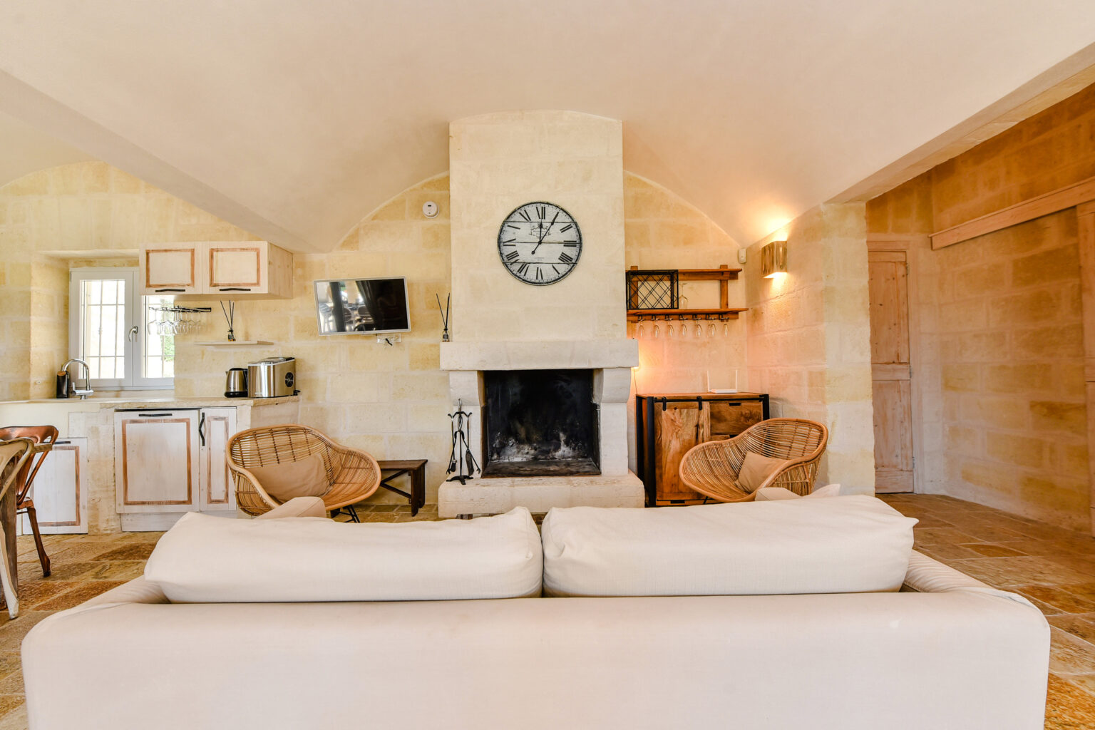 Cozy living area featuring a stone fireplace, modern kitchen with wooden cabinets, and stylish rattan chairs. The space is illuminated by natural light and has a neutral color palette, creating a warm and inviting atmosphere. | Only on SopranoVillas