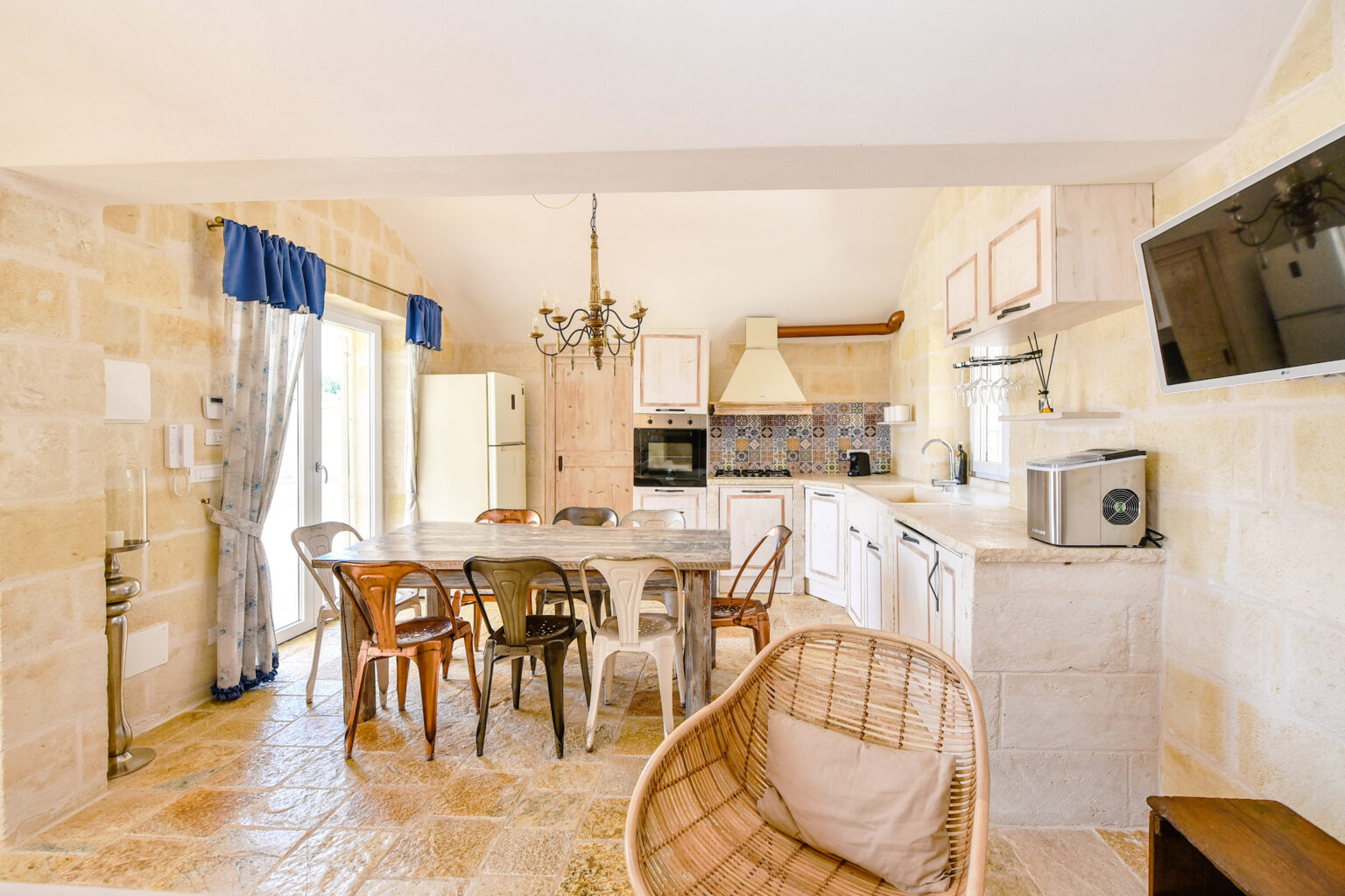 Bright and airy kitchen interior featuring a large dining table surrounded by wooden chairs, stone walls, and modern appliances. Natural light floods the space through large windows, complemented by decorative curtains and a chandelier. Ideal for family gatherings and cooking enthusiasts. | Only on SopranoVillas
