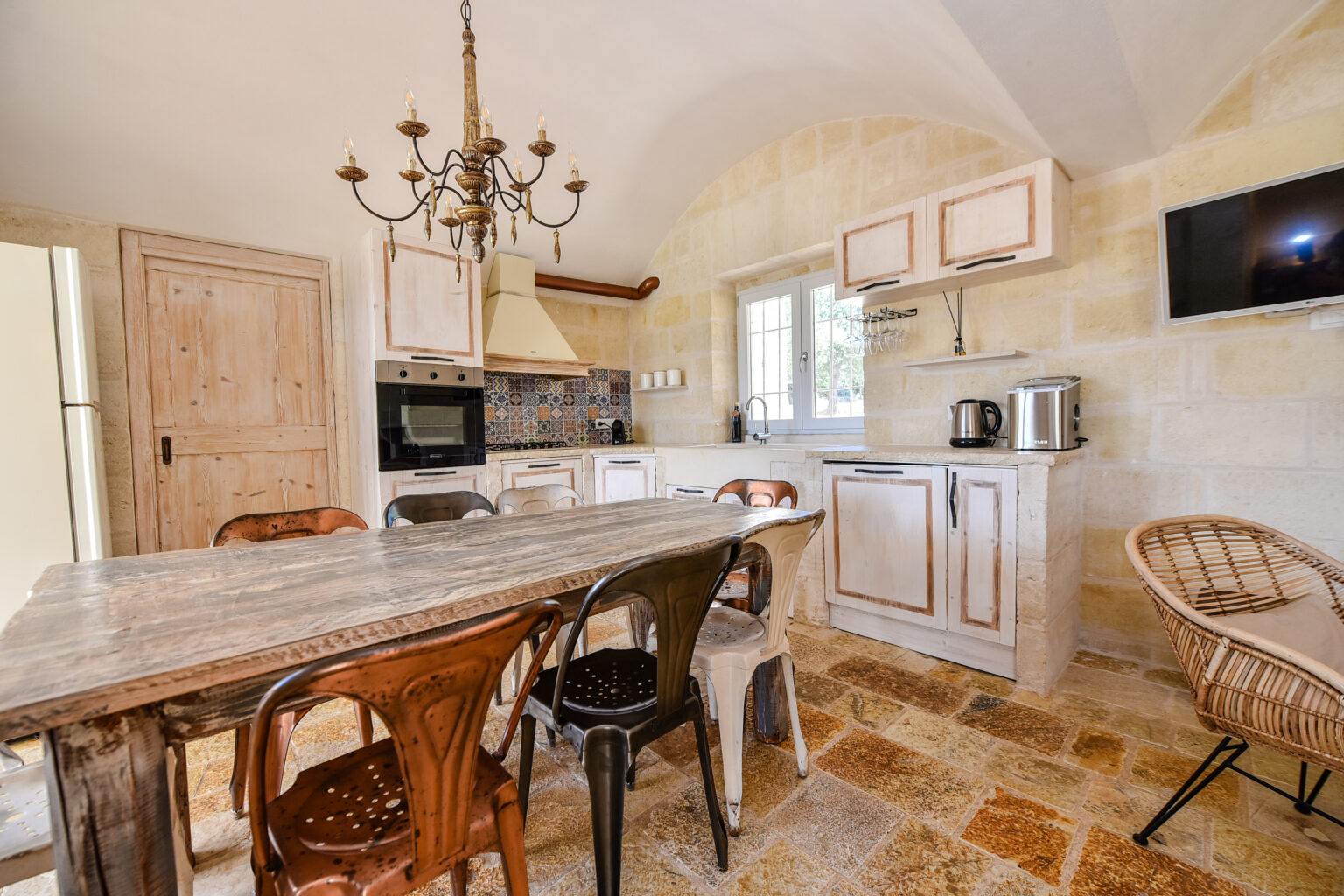 Spacious kitchen featuring a rustic wooden dining table surrounded by metal chairs, a stylish chandelier, modern appliances, and natural stone walls, creating a warm and inviting atmosphere. | Only on SopranoVillas