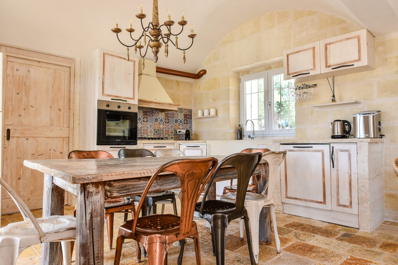 Modern kitchen interior featuring a wooden dining table surrounded by vintage metal chairs, a rustic chandelier, and a stylish backsplash. The kitchen includes built-in appliances, a window providing natural light, and a warm stone floor, creating a cozy and inviting atmosphere. | Only on SopranoVillas
