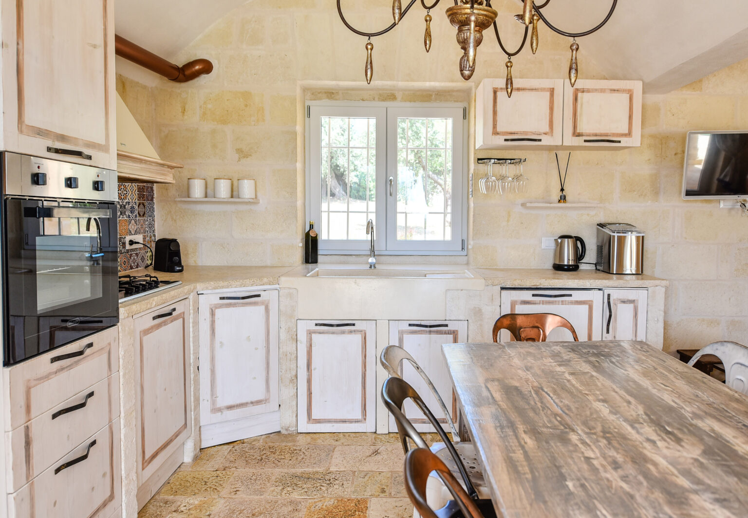 Modern kitchen interior featuring light-colored wooden cabinets, a spacious dining table, and stainless steel appliances, with natural light streaming through a window. | Only on SopranoVillas