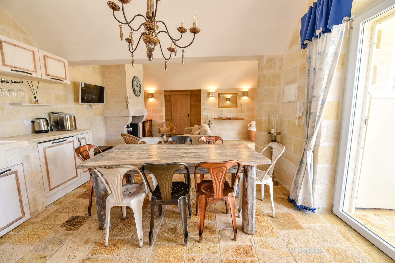 Spacious kitchen dining area featuring a large wooden table surrounded by colorful chairs, a stylish chandelier, and natural light from large windows, showcasing a warm and inviting atmosphere. | Only on SopranoVillas