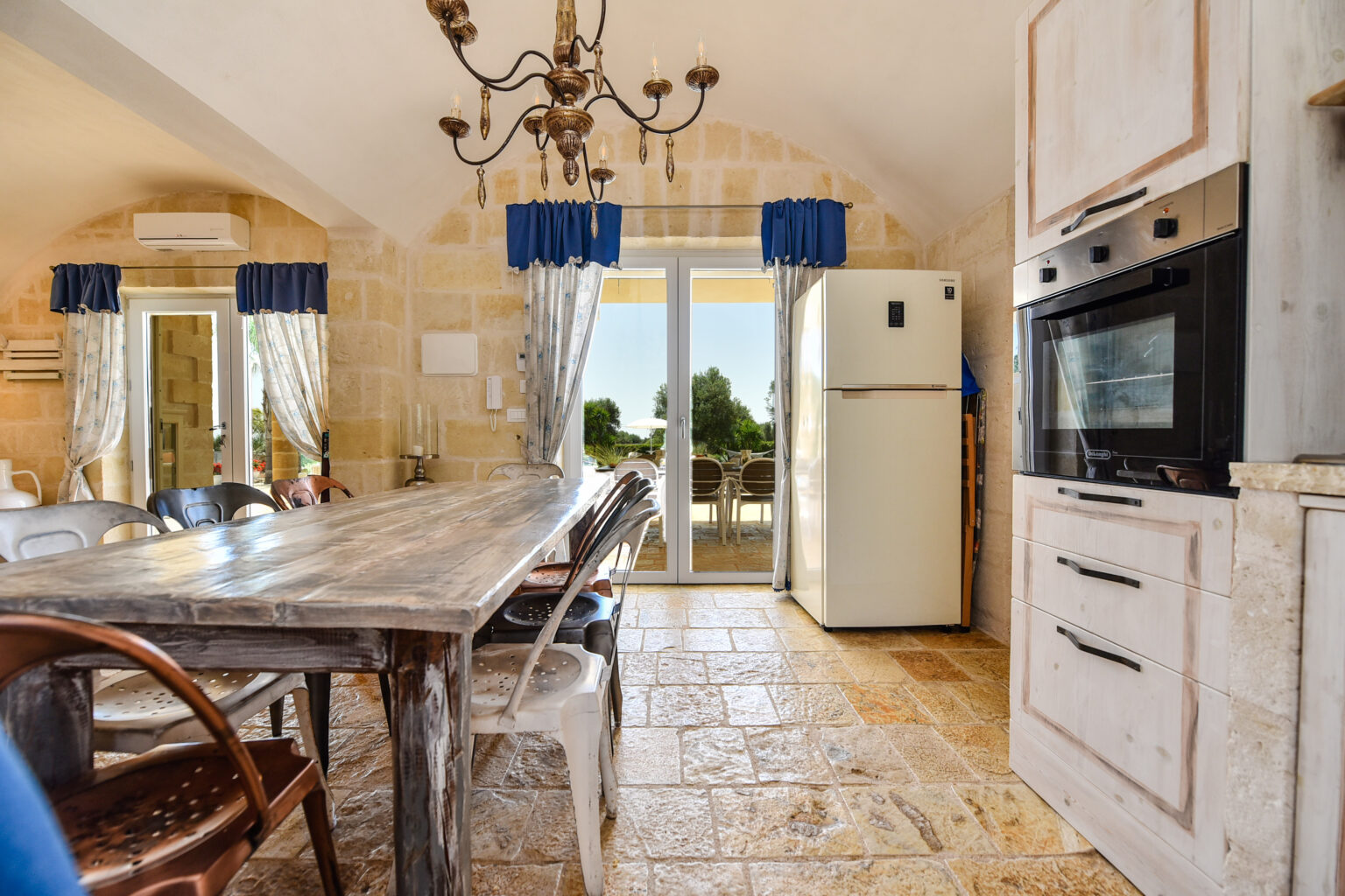 Spacious kitchen with a rustic wooden dining table, white appliances, and stone walls, featuring large windows with blue curtains that offer a view of the outdoor area. | Only on SopranoVillas