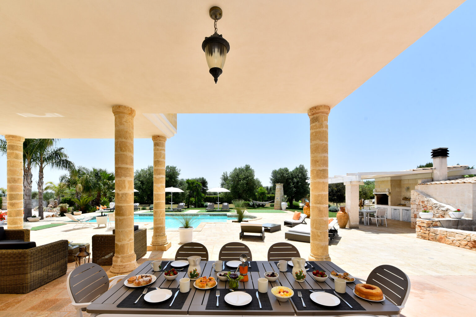 Dining area with a beautifully set table featuring various dishes, overlooking a luxurious outdoor pool and landscaped garden, framed by elegant stone columns and clear blue skies. | Only on SopranoVillas