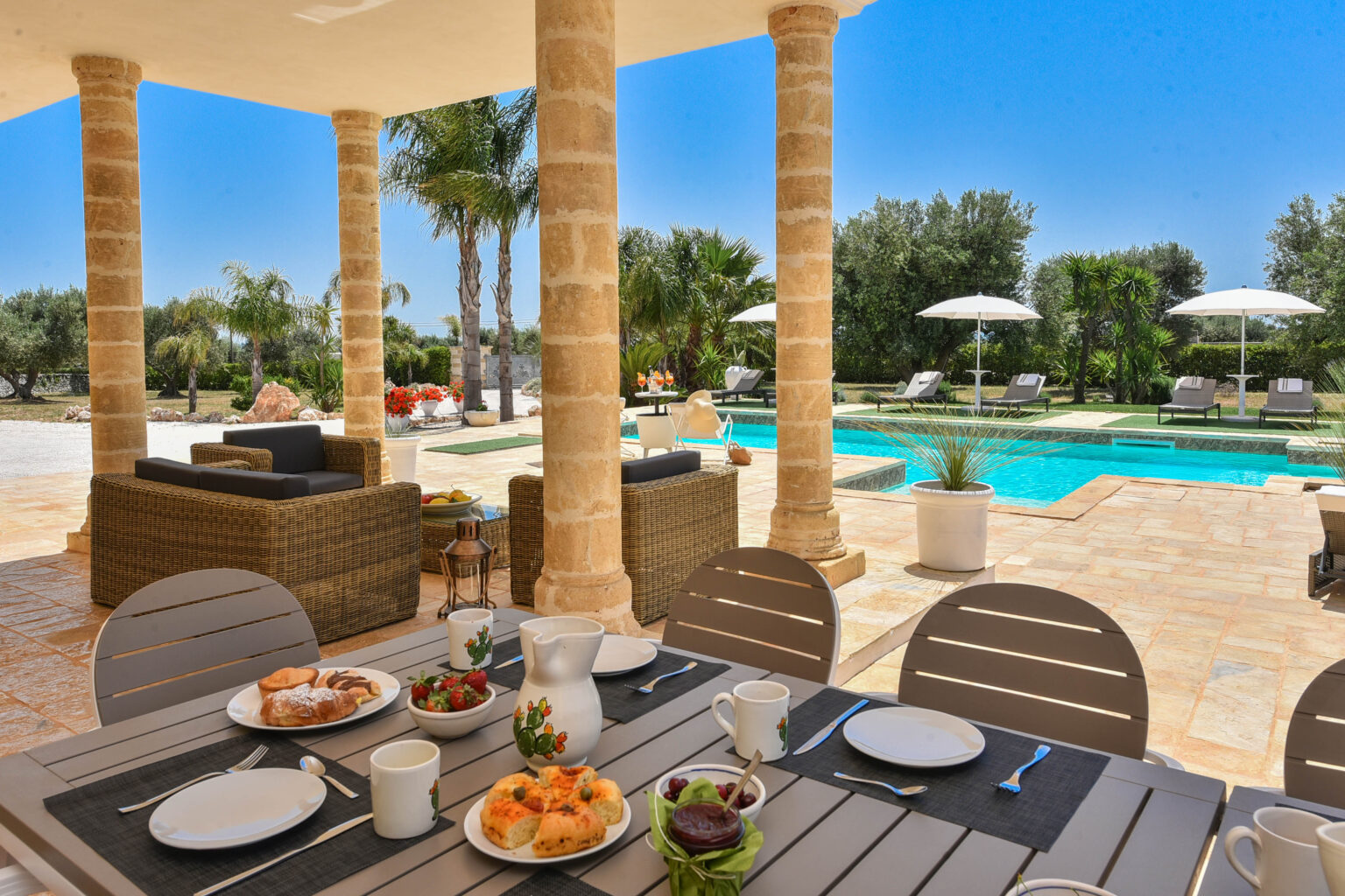 Outdoor dining area with a beautifully set table featuring breakfast dishes, overlooking a luxurious pool surrounded by palm trees and lounge chairs under umbrellas. | Only on SopranoVillas