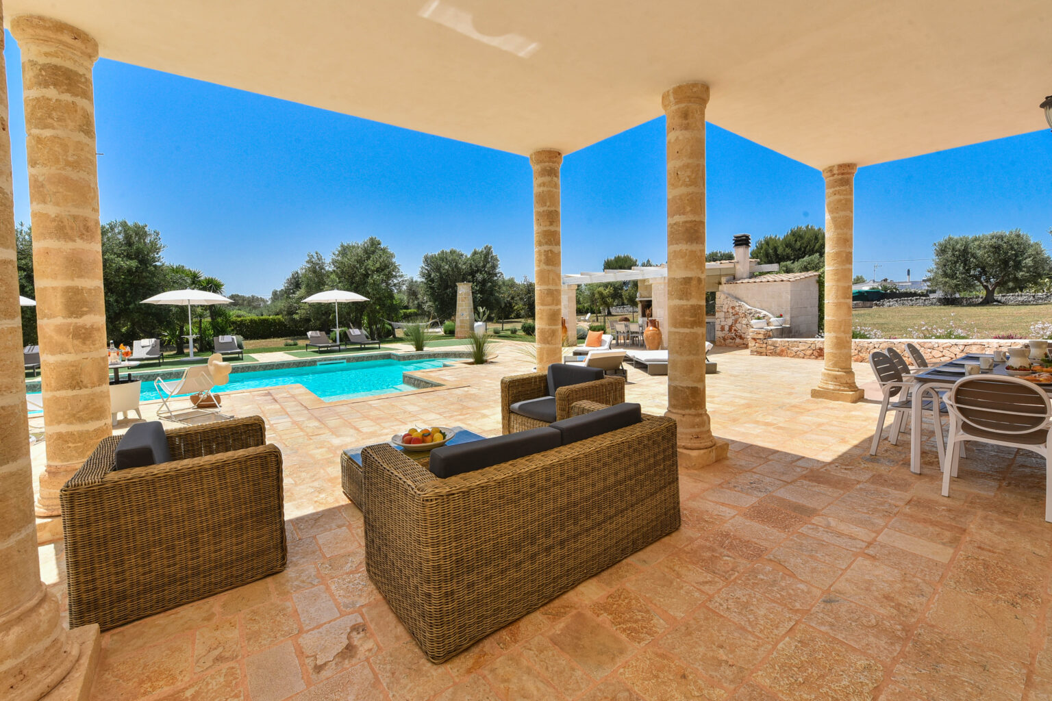 Modern outdoor patio with comfortable seating, overlooking a swimming pool surrounded by lush greenery and sun loungers. Bright blue sky enhances the relaxing atmosphere, perfect for outdoor gatherings. | Only on SopranoVillas
