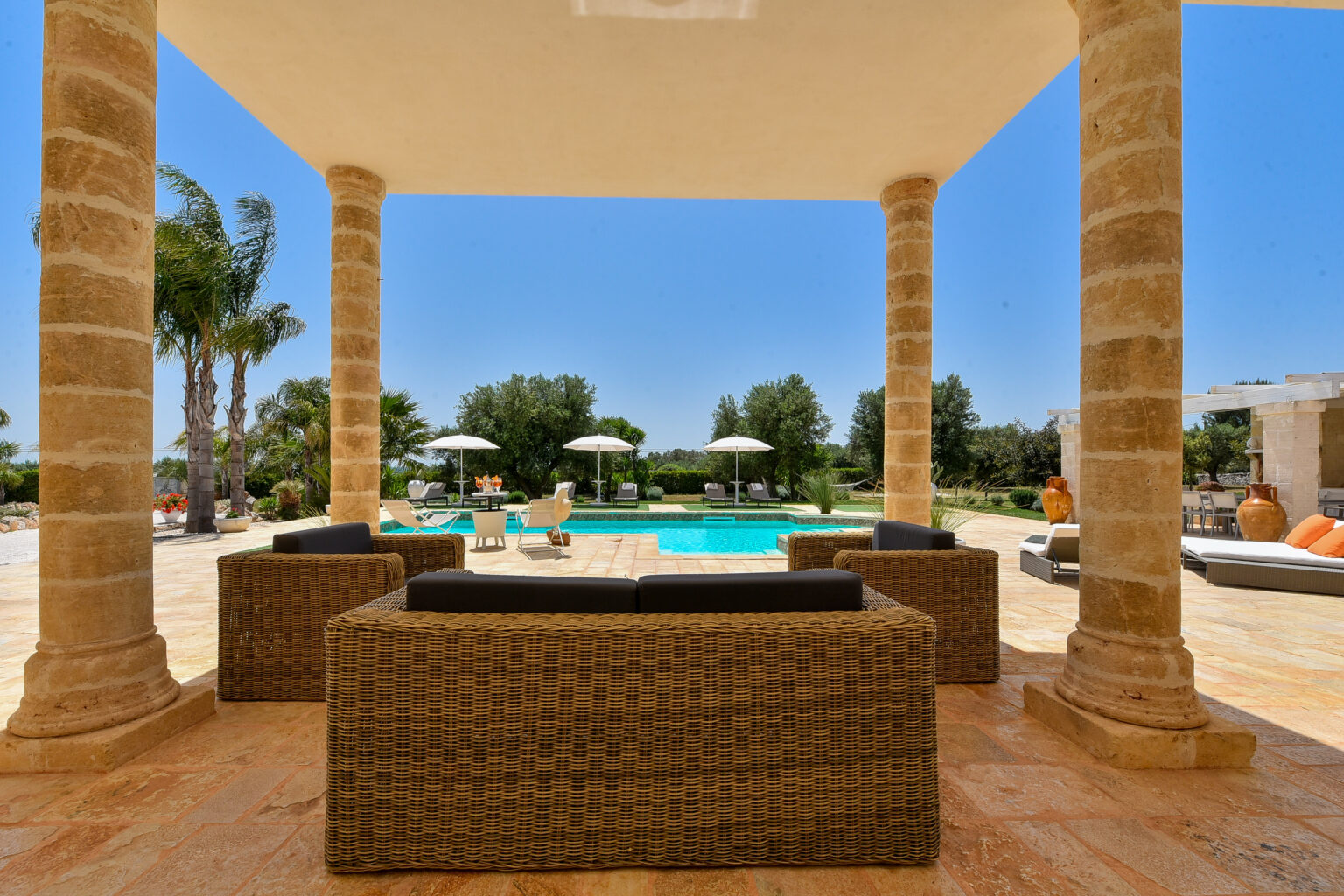 Luxurious outdoor living area featuring wicker seating, stone columns, and a view of a sparkling pool surrounded by lush greenery and sun loungers under umbrellas. Ideal for relaxation and entertaining in a beautiful, sunny setting. | Only on SopranoVillas