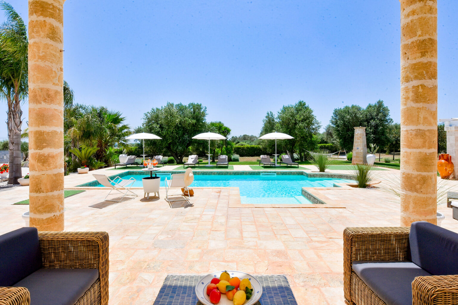Luxurious outdoor pool area featuring a sparkling blue pool, lounge chairs with umbrellas, and a scenic garden backdrop. A cozy seating arrangement with a plate of fresh fruit enhances the inviting atmosphere under a clear blue sky. | Only on SopranoVillas