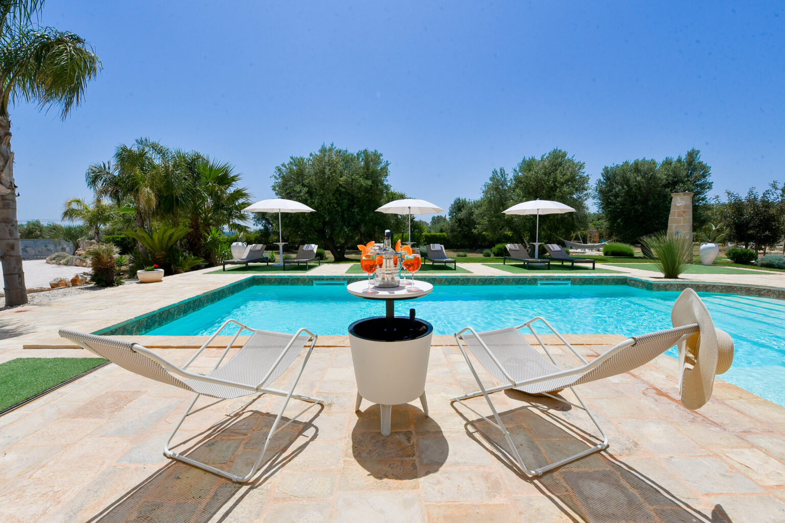 Luxurious outdoor pool area featuring two stylish lounge chairs, a table with refreshing cocktails, and elegant umbrellas, surrounded by lush greenery under a clear blue sky. Perfect for relaxation and leisure in a sunny setting. | Only on SopranoVillas
