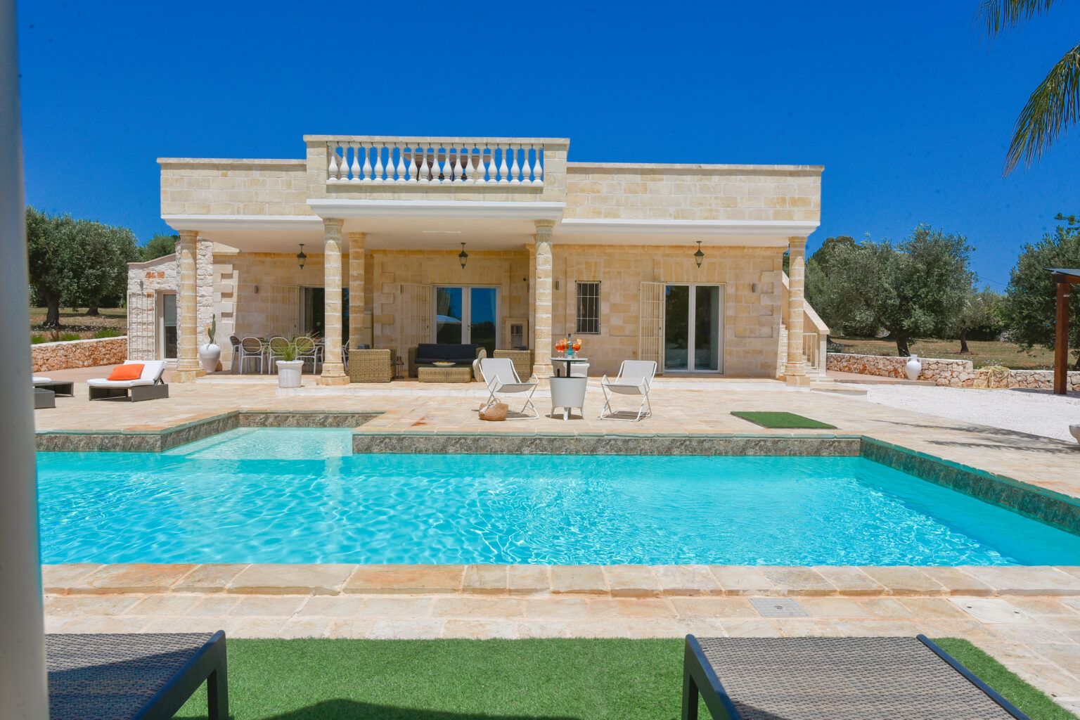 Luxurious stone villa with a swimming pool, surrounded by olive trees under a clear blue sky. Features a spacious terrace with lounge chairs and outdoor seating. | Only on SopranoVillas