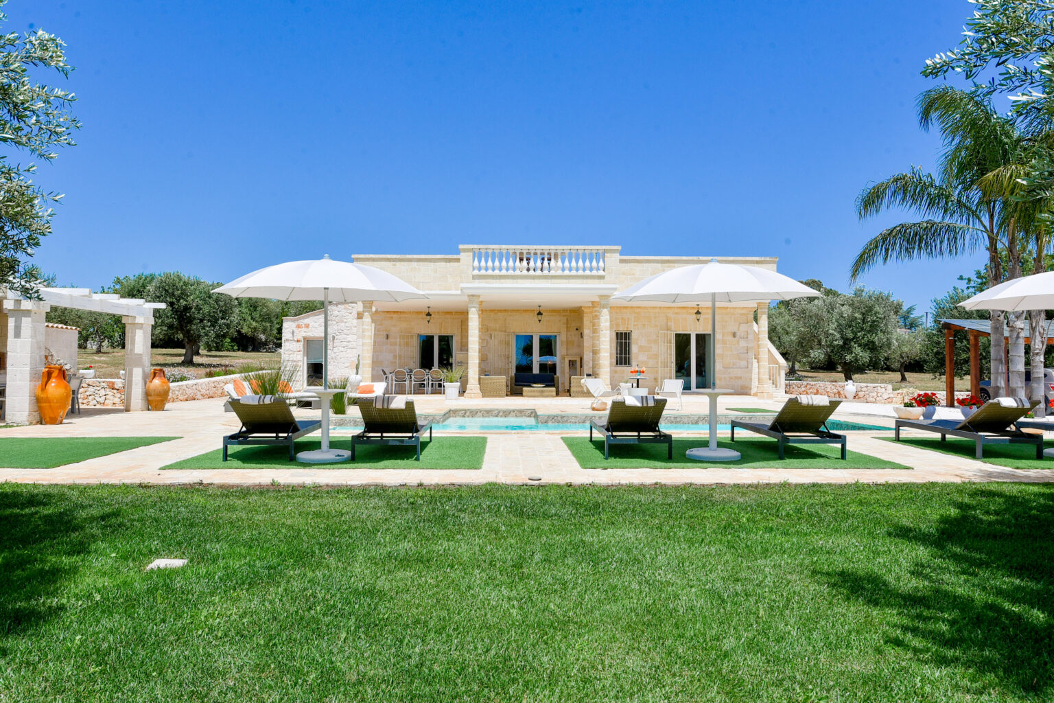 Luxurious villa with a swimming pool, surrounded by green lawns and palm trees, featuring sun loungers and shaded seating areas, ideal for relaxation in a sunny outdoor setting. | Only on SopranoVillas