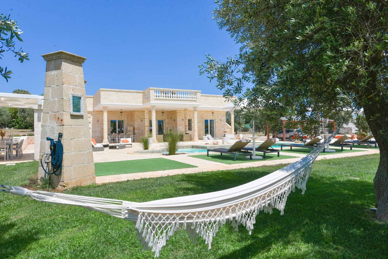 Serene outdoor view of a luxury villa featuring a spacious garden, a hammock, and sun loungers under clear blue skies, ideal for relaxation and outdoor living. | Only on SopranoVillas