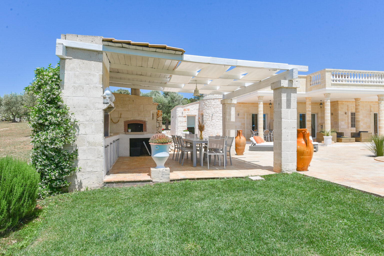 Outdoor patio area featuring a stone structure with a shaded dining space, barbecue grill, and terracotta pots, surrounded by lush green grass and a clear blue sky. | Only on SopranoVillas