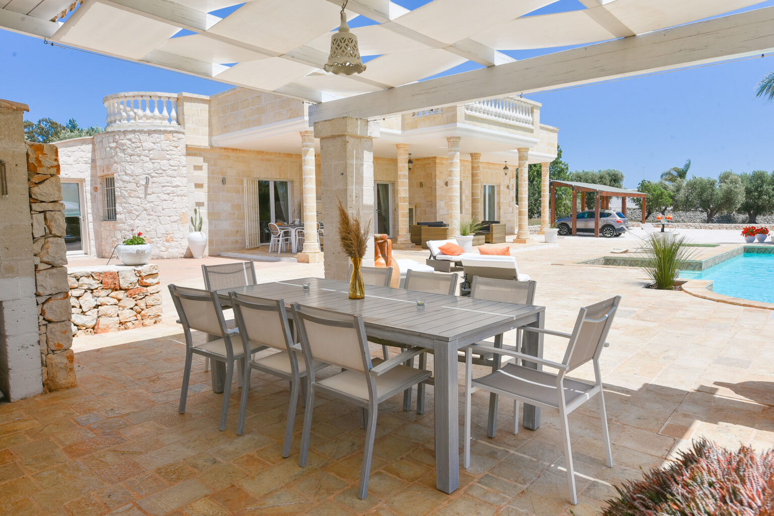 Outdoor dining area with a modern table and chairs under a pergola, set beside a luxurious pool and a stone villa surrounded by greenery. Perfect for entertaining in a sunny, Mediterranean setting. | Only on SopranoVillas