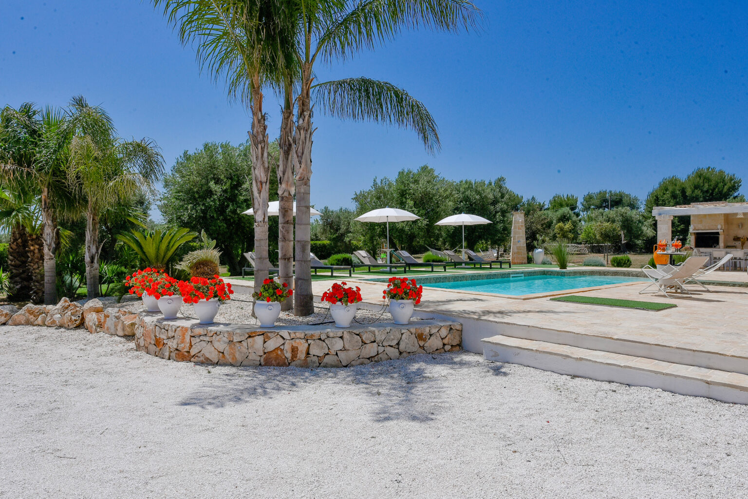 Scenic outdoor pool area with palm trees, vibrant flower pots, and sun loungers under white umbrellas, surrounded by lush greenery and a clear blue sky. Ideal for relaxation and summer gatherings. | Only on SopranoVillas