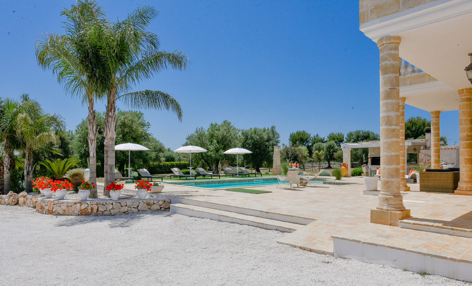 Stunning outdoor pool area featuring lush palm trees, vibrant flowers, and sun loungers under white umbrellas, set against a clear blue sky. Ideal for relaxation and outdoor entertaining. | Only on SopranoVillas