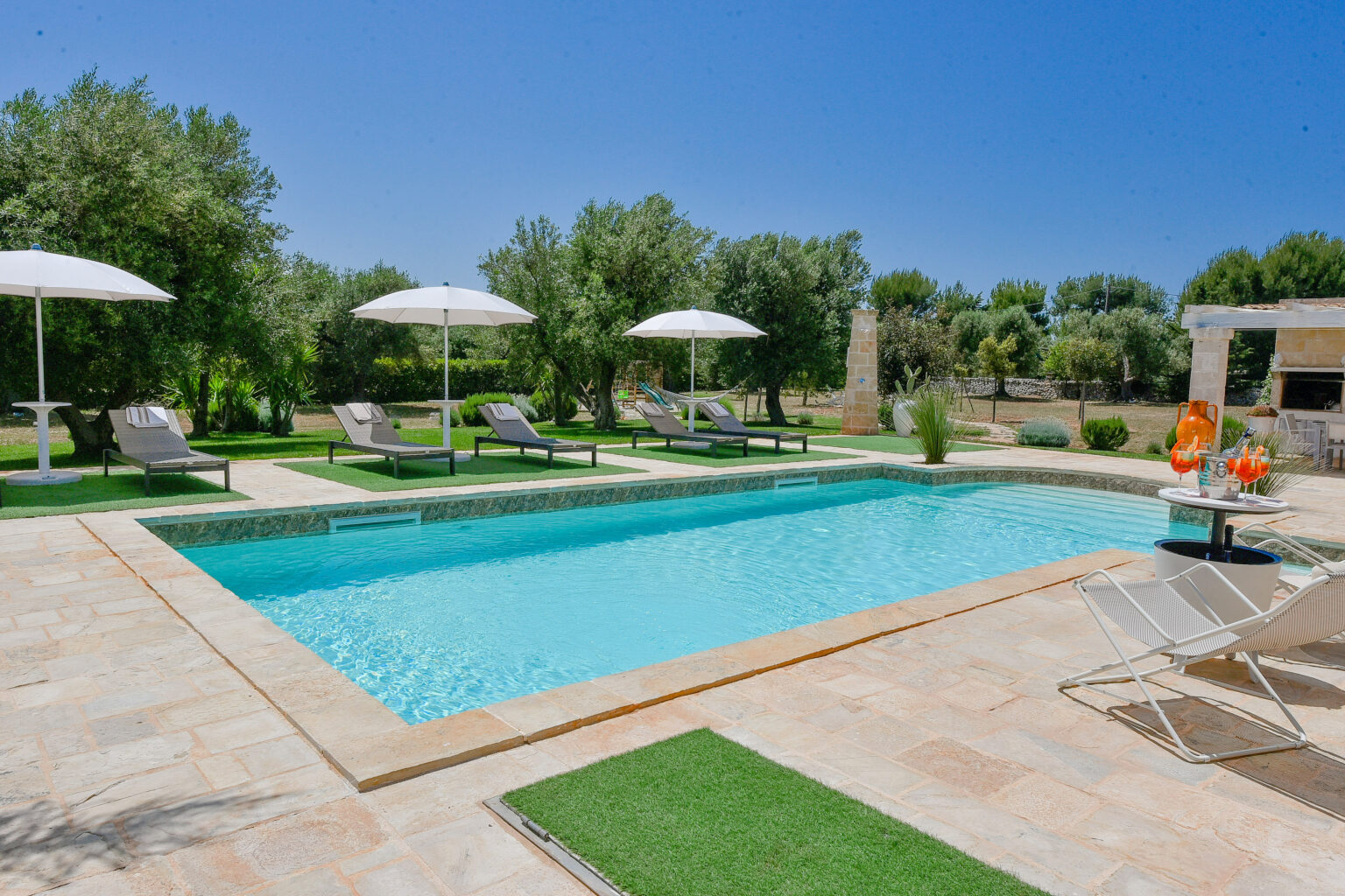 Luxurious outdoor pool area featuring a sparkling blue swimming pool, comfortable lounge chairs, and shaded umbrellas, surrounded by lush greenery and a well-maintained garden. Perfect for relaxation and leisure. | Only on SopranoVillas