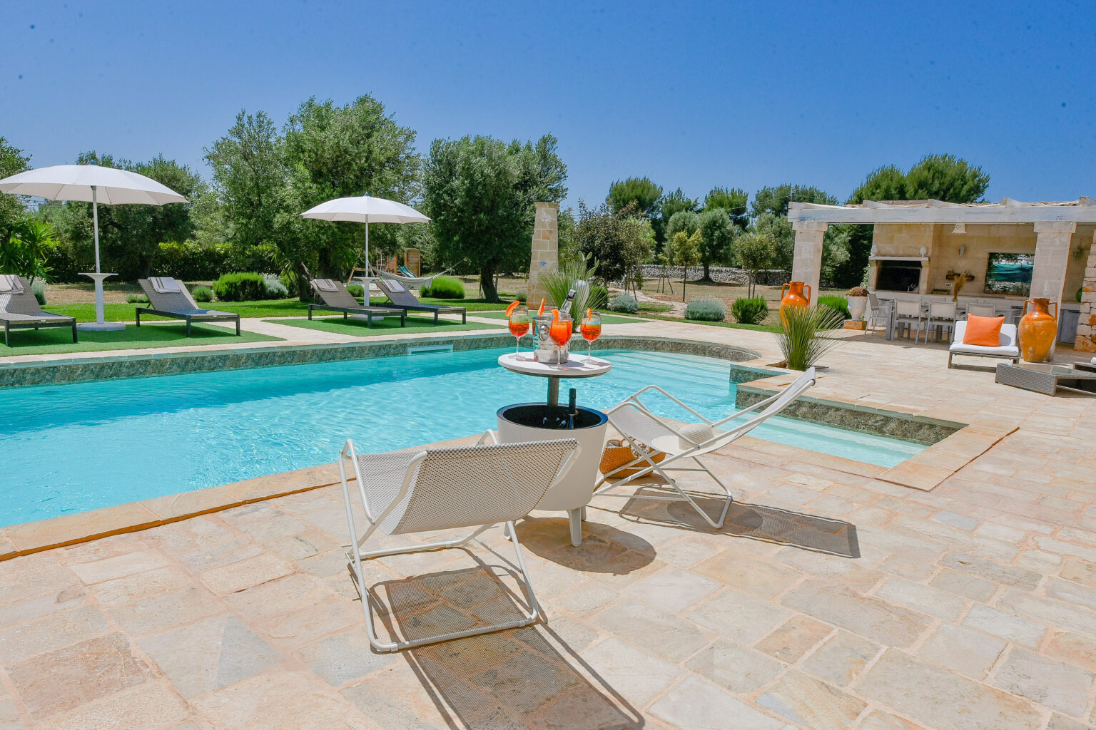 Luxurious outdoor pool area featuring sun loungers and umbrellas, surrounded by lush greenery, with a stylish table set for drinks, ideal for relaxation and leisure. | Only on SopranoVillas