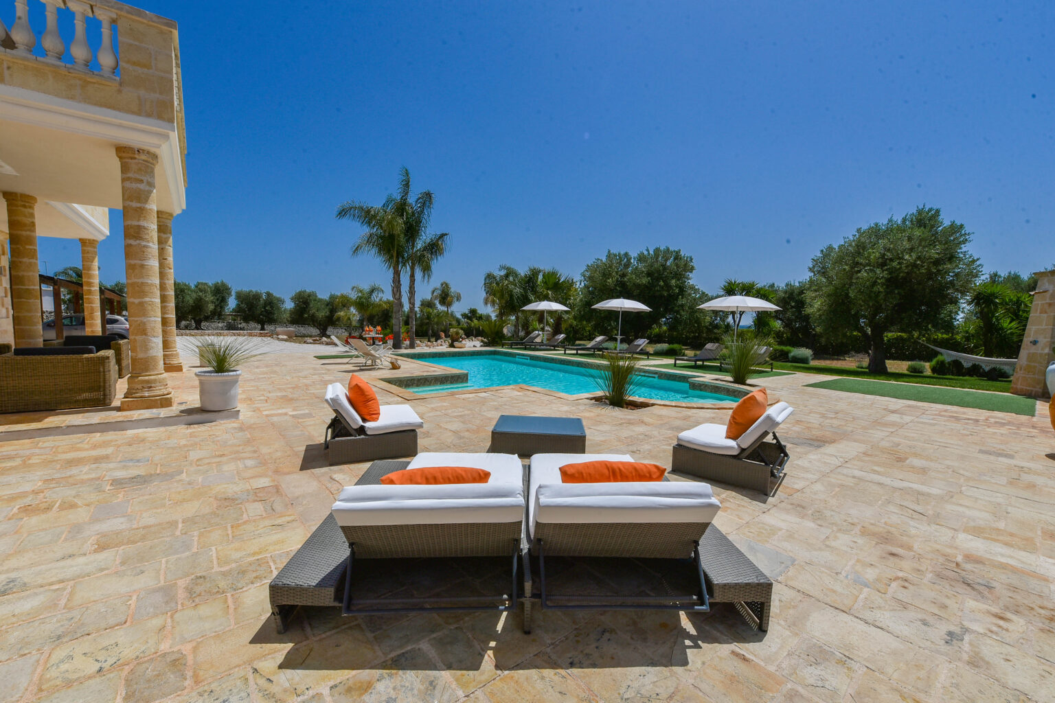 Luxurious outdoor pool area featuring modern sun loungers with orange cushions, surrounded by lush greenery and palm trees under a clear blue sky. Perfect for relaxation and leisure. | Only on SopranoVillas
