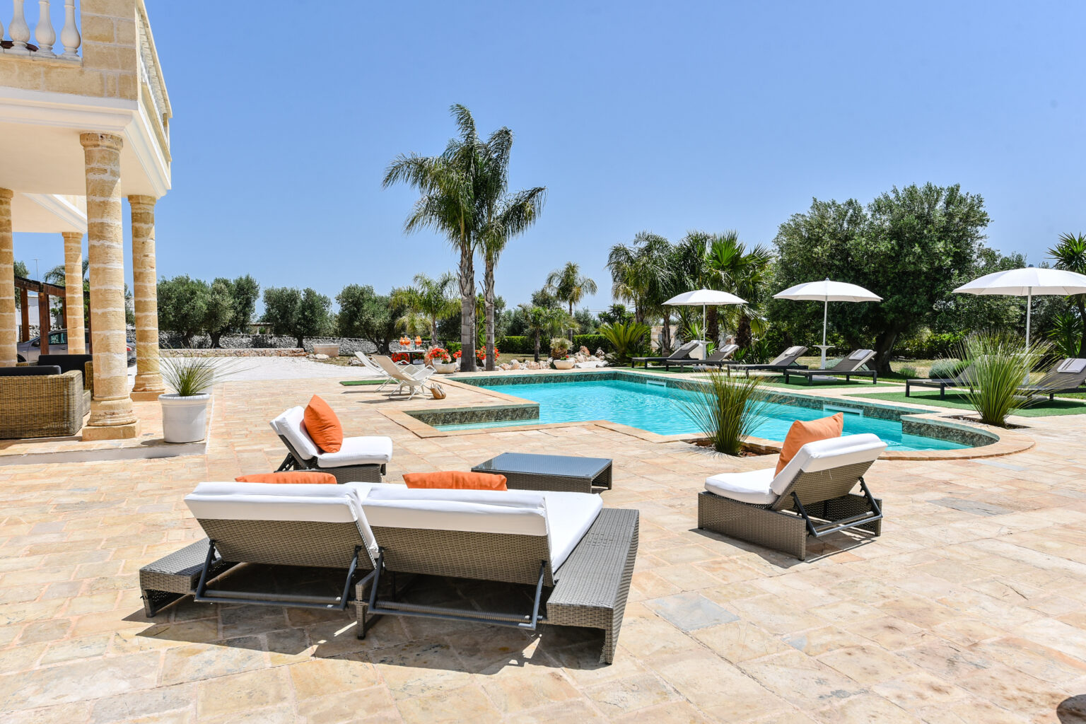 Luxurious outdoor pool area featuring comfortable lounge chairs with orange cushions, surrounded by palm trees and sun umbrellas, set against a clear blue sky and a stone patio. Perfect for relaxation and leisure. | Only on SopranoVillas