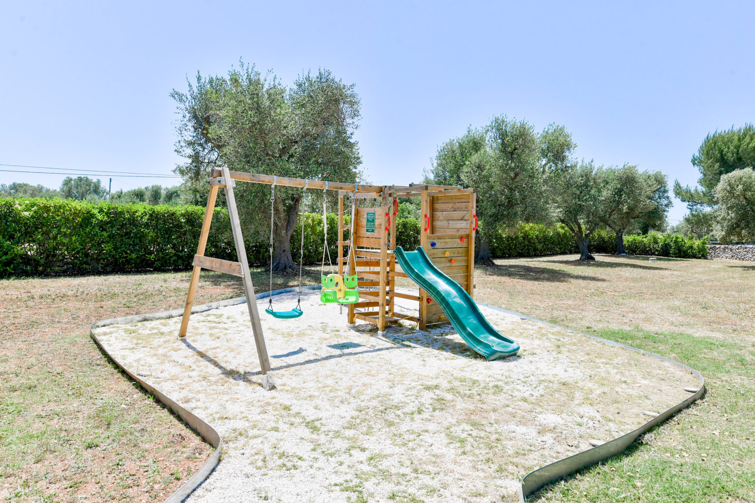 Wooden playground set featuring a slide, swings, and climbing wall, situated in a grassy outdoor area surrounded by olive trees. Ideal for children's recreational activities in a sunny environment. | Only on SopranoVillas