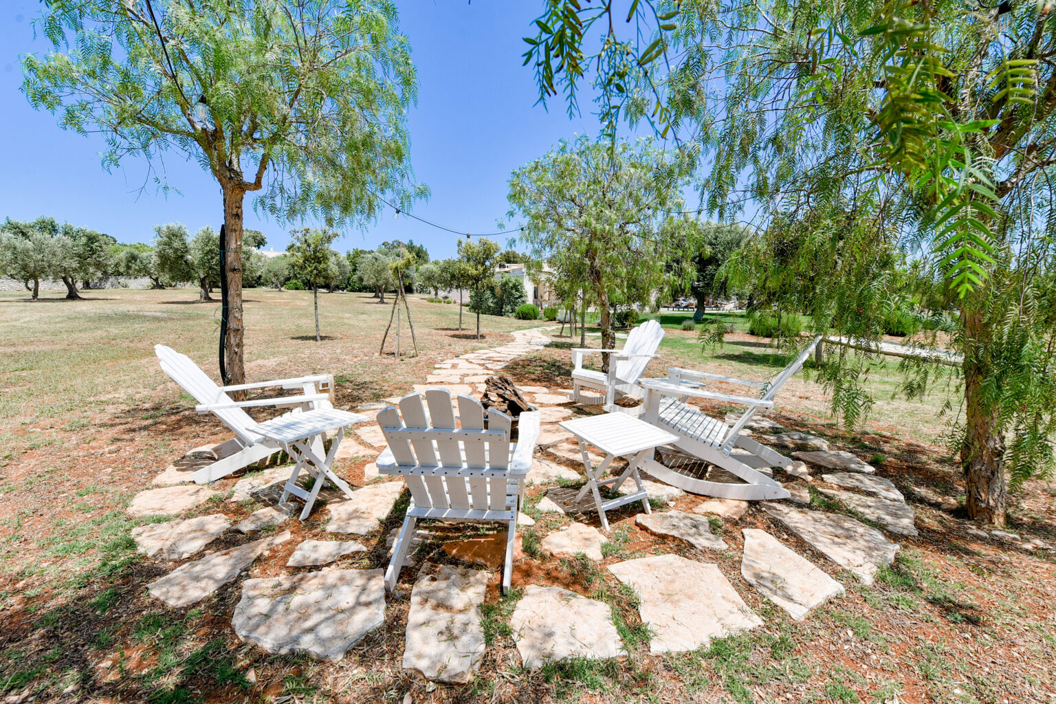 Cozy outdoor seating area with white Adirondack chairs arranged around a stone fire pit, surrounded by trees and a grassy landscape, ideal for relaxation and gatherings. | Only on SopranoVillas