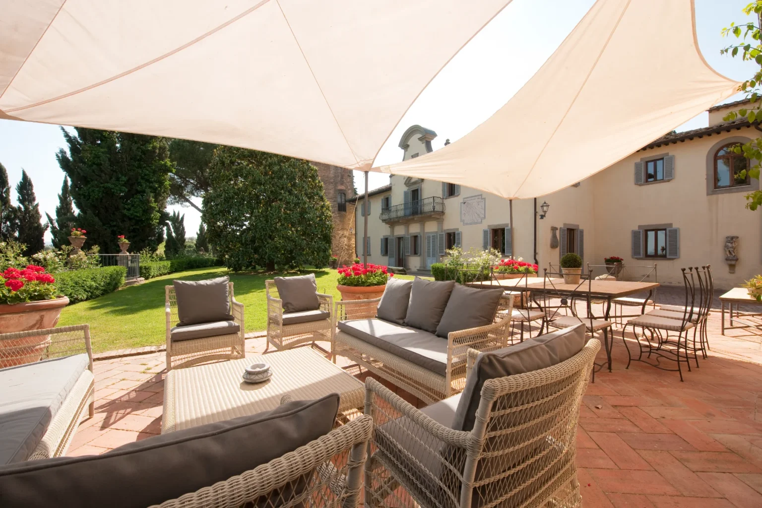Outdoor seating area with comfortable wicker furniture and gray cushions, shaded by a large fabric canopy, overlooking a lush garden and a rustic villa in the background. Perfect for relaxation and social gatherings. | Only on SopranoVillas