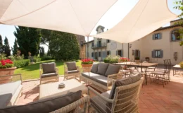 Outdoor seating area with comfortable wicker furniture and gray cushions, shaded by a large fabric canopy, overlooking a lush garden and a rustic villa in the background. Perfect for relaxation and social gatherings.