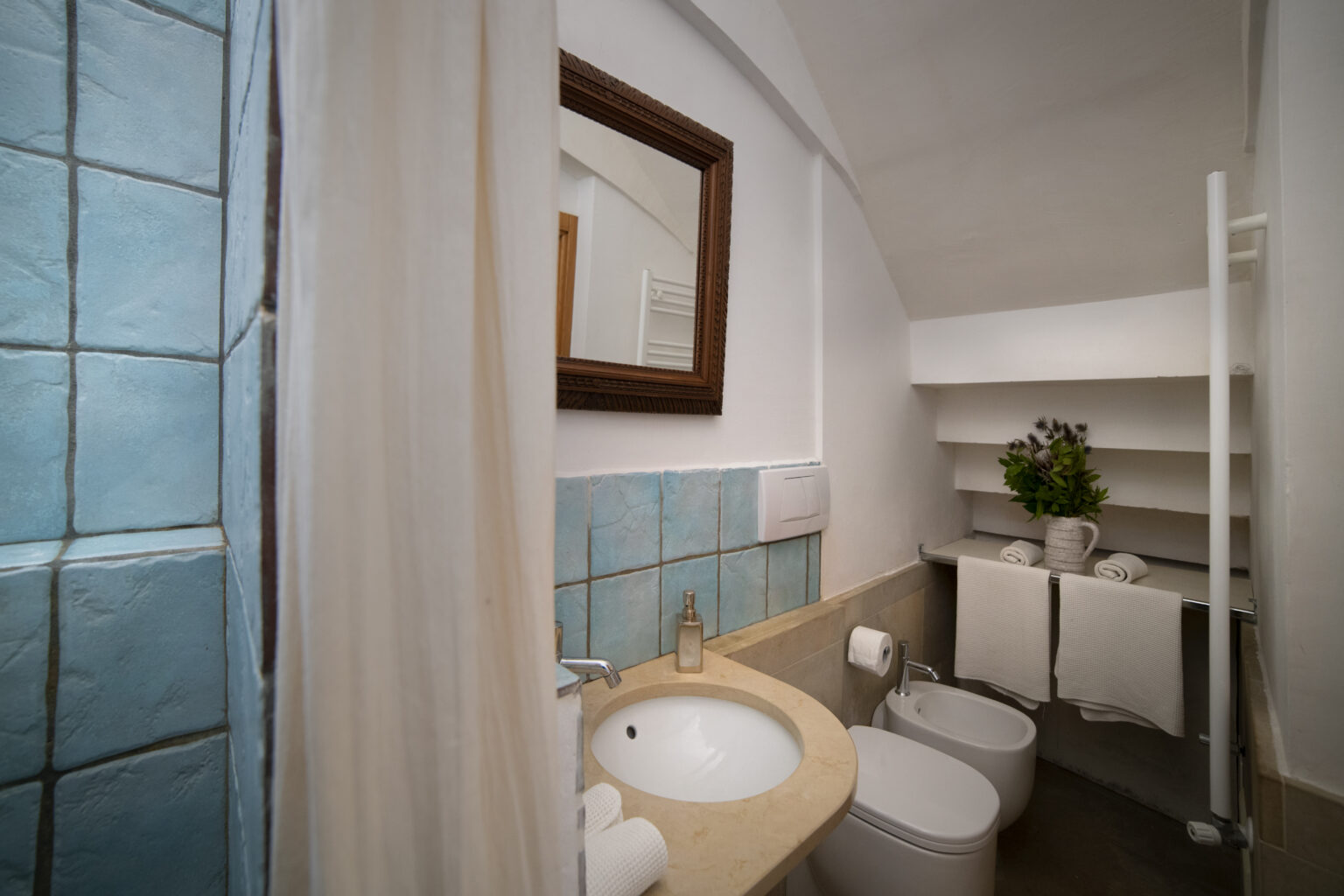 Modern bathroom featuring turquoise tiled walls, a round sink with a marble countertop, a mirror, and neatly arranged towels. The space includes a bidet, toilet, and a decorative plant, emphasizing a clean and inviting atmosphere. Ideal for showcasing interior design in home decor or rental listings. | Only on SopranoVillas