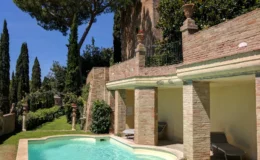 Scenic outdoor pool surrounded by lush greenery and elegant brick architecture, with tall trees and a clear blue sky in the background, ideal for relaxation and leisure activities.