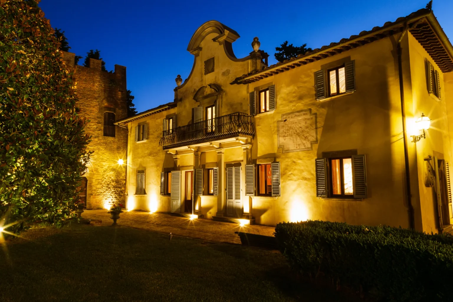 Historic villa illuminated at night with a lush garden, showcasing architectural details and a stone tower in the background. | Only on SopranoVillas