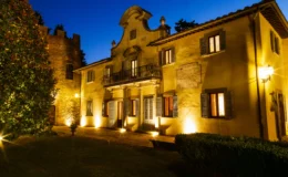 Historic villa illuminated at night with a lush garden, showcasing architectural details and a stone tower in the background.