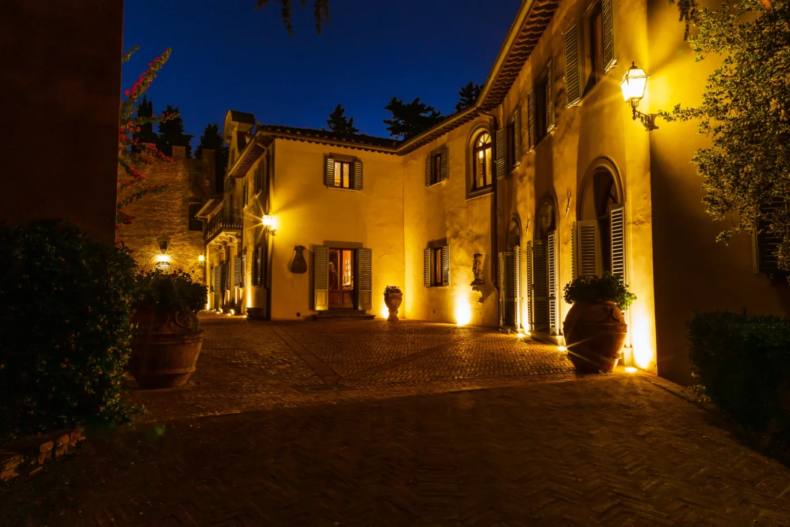 Elegant villa entrance illuminated at night, featuring warm lighting, lush greenery, and decorative pots. The cobblestone driveway leads to a charming facade with arched windows and shutters, surrounded by trees. Ideal for showcasing luxury architecture and outdoor ambiance. | Only on SopranoVillas