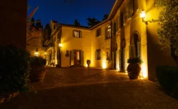 Elegant villa entrance illuminated at night, featuring warm lighting, lush greenery, and decorative pots. The cobblestone driveway leads to a charming facade with arched windows and shutters, surrounded by trees. Ideal for showcasing luxury architecture and outdoor ambiance.