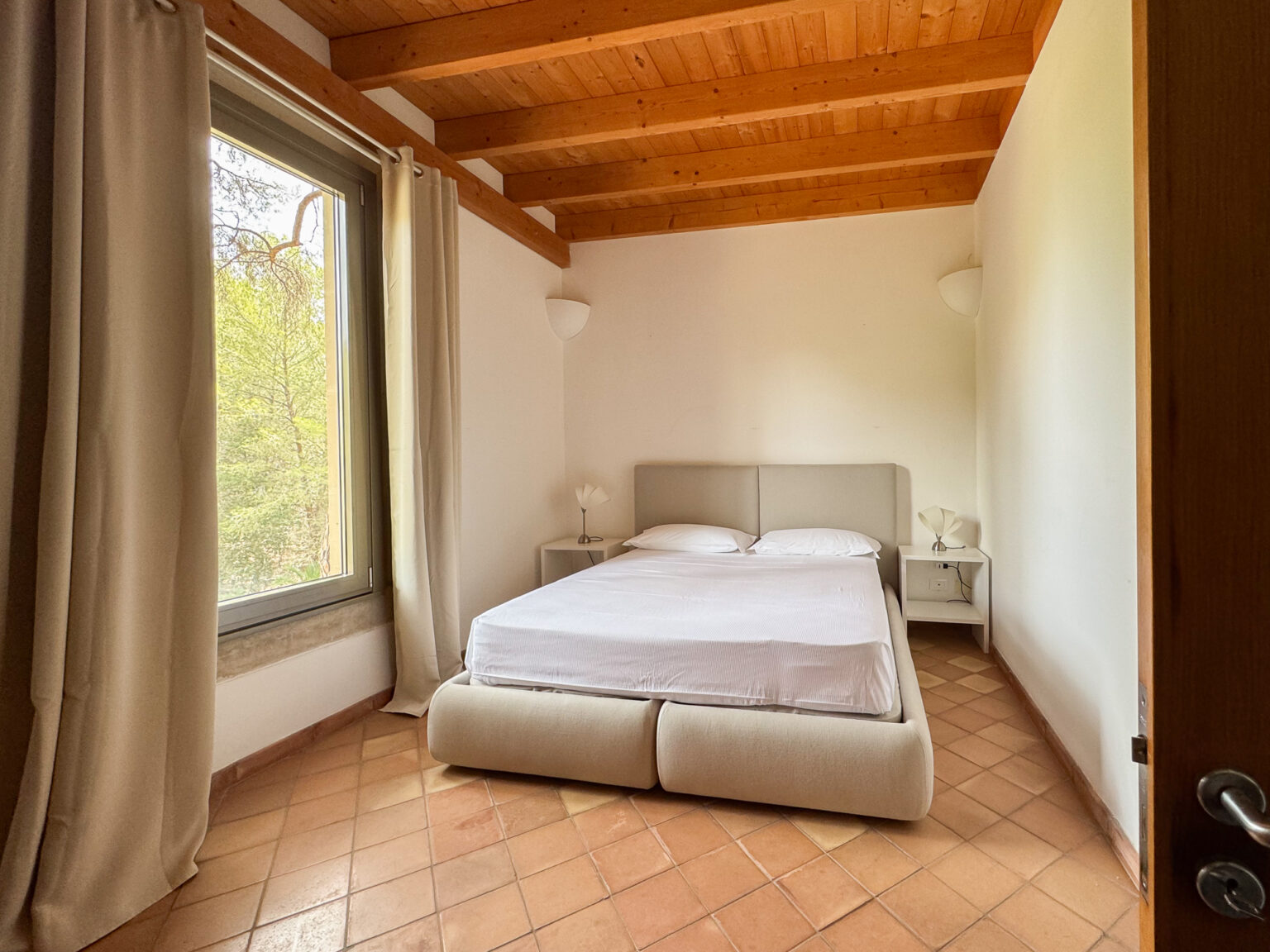 Cozy bedroom featuring a double bed with a light-colored upholstered headboard, minimalist nightstand with a lamp, and large window with natural light illuminating the space, showcasing a serene outdoor view. The room has wooden ceiling beams and terracotta tile flooring, creating a warm and inviting atmosphere. | Only on SopranoVillas