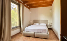 Cozy bedroom featuring a double bed with a light-colored upholstered headboard, minimalist nightstand with a lamp, and large window with natural light illuminating the space, showcasing a serene outdoor view. The room has wooden ceiling beams and terracotta tile flooring, creating a warm and inviting atmosphere.