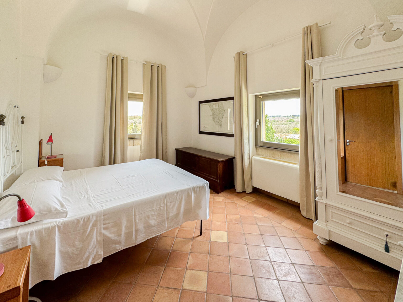 Bright and airy bedroom featuring a white bed with a neatly arranged bedspread, a bedside lamp, and a wooden dresser. Natural light streams through a window adorned with beige curtains, illuminating the terracotta tile floor and white walls. A vintage wardrobe and a framed map add character to the room, creating a cozy and inviting atmosphere. | Only on SopranoVillas