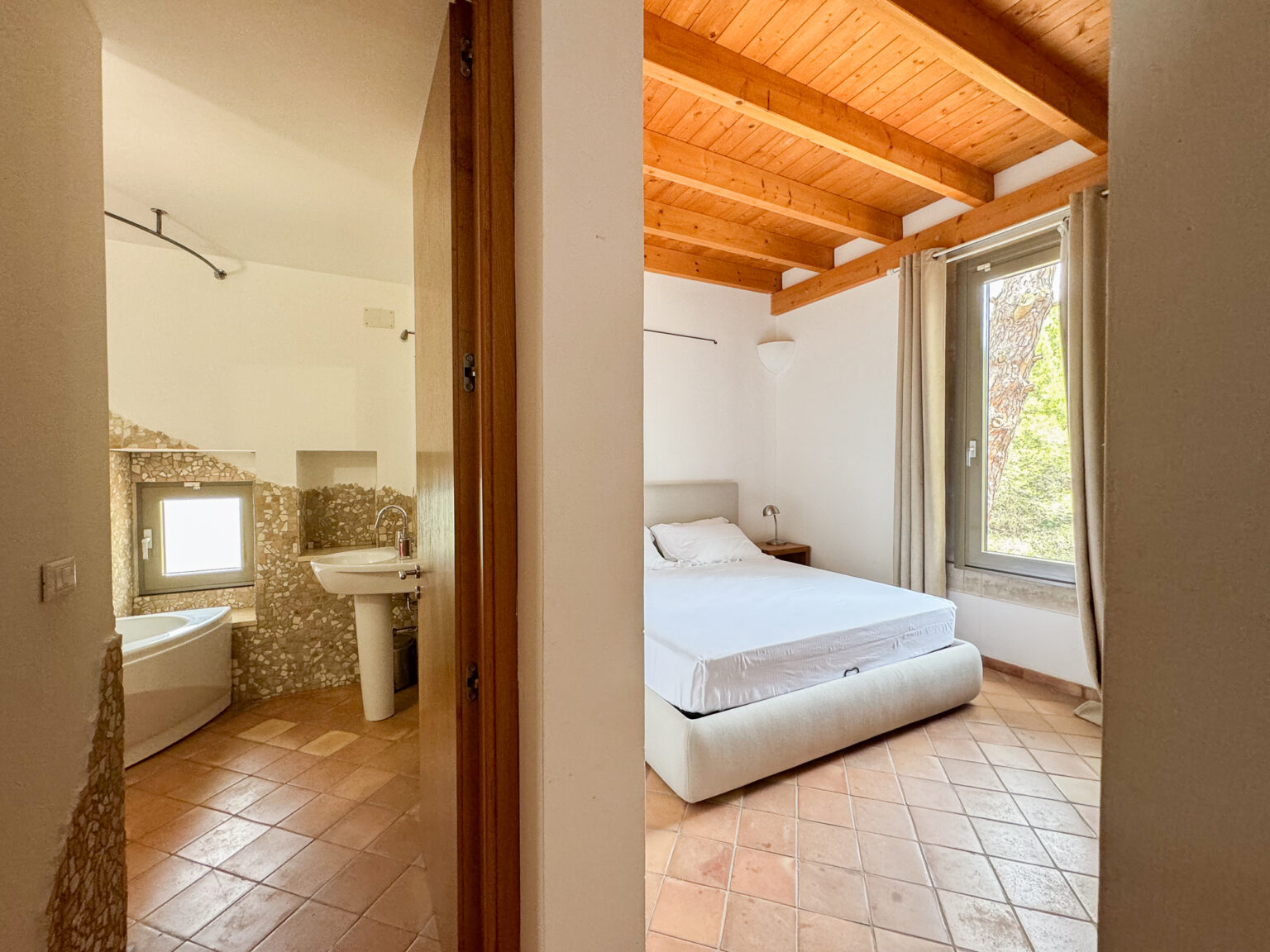 Spacious interior view of a modern bedroom with a double bed and natural light from a large window, adjacent to a stylish bathroom featuring a bathtub and sink, showcasing terracotta flooring and wooden ceiling beams. | Only on SopranoVillas